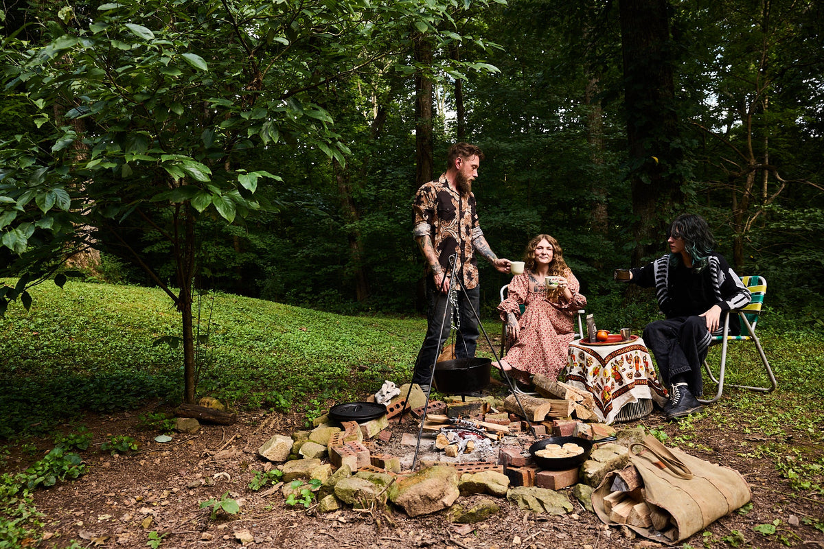 Three people gathered outdoors in a wooded campsite setting, with a person standing near a hanging pot, a seated woman in the center, and another person at a small table with a patterned blanket. 