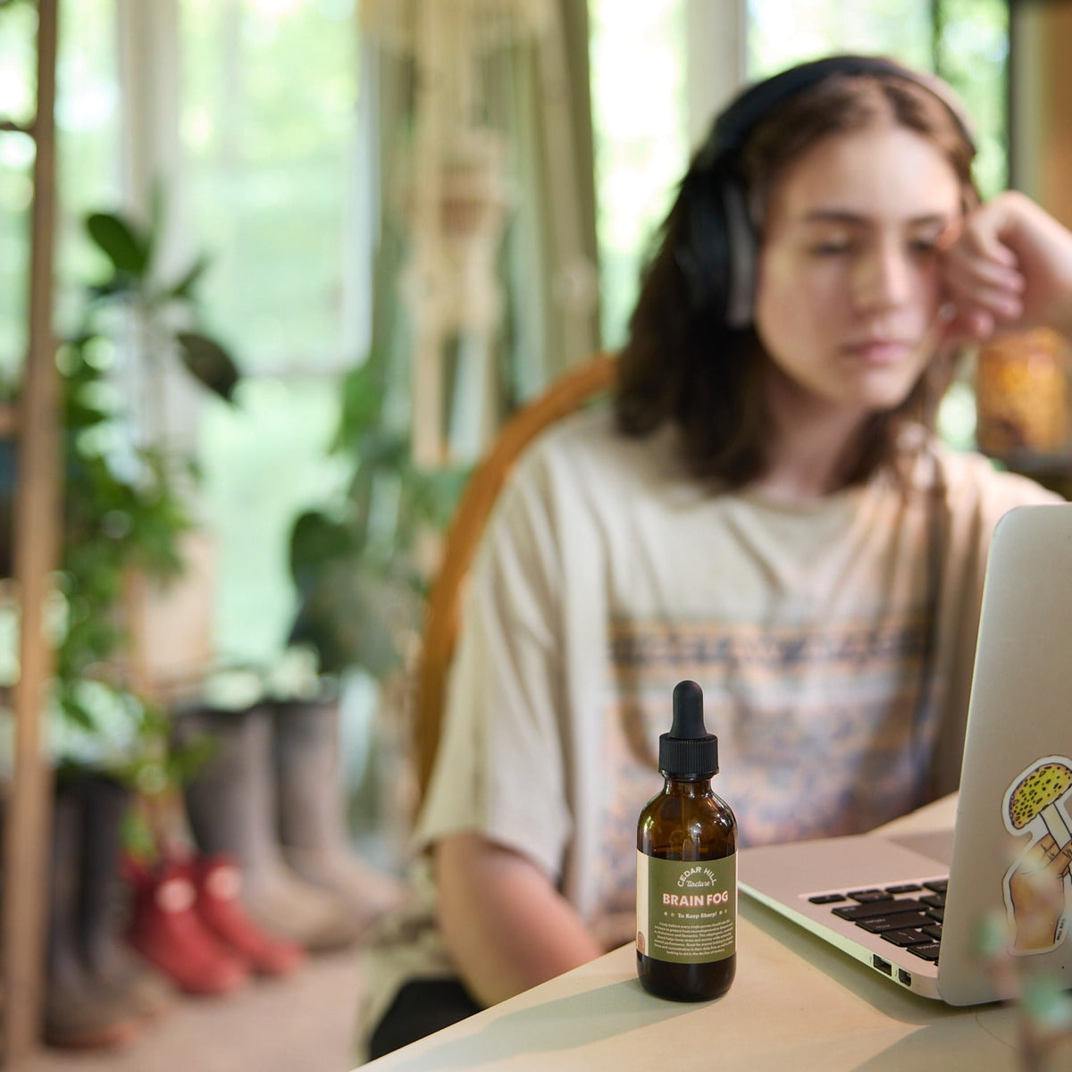 A student wearing headphones uses a laptop near a bottle of Cedar Hill Brain Fog tincture.
