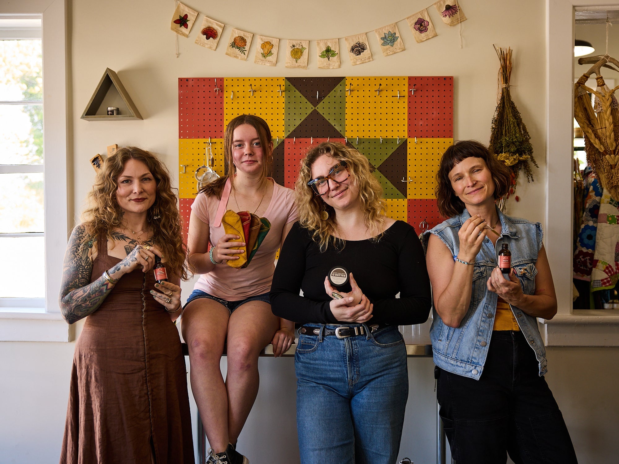 Four women smiling and holding Cedar Hill Homestead herbal products inside the store, with dried herbs and a botanical banner overhead.