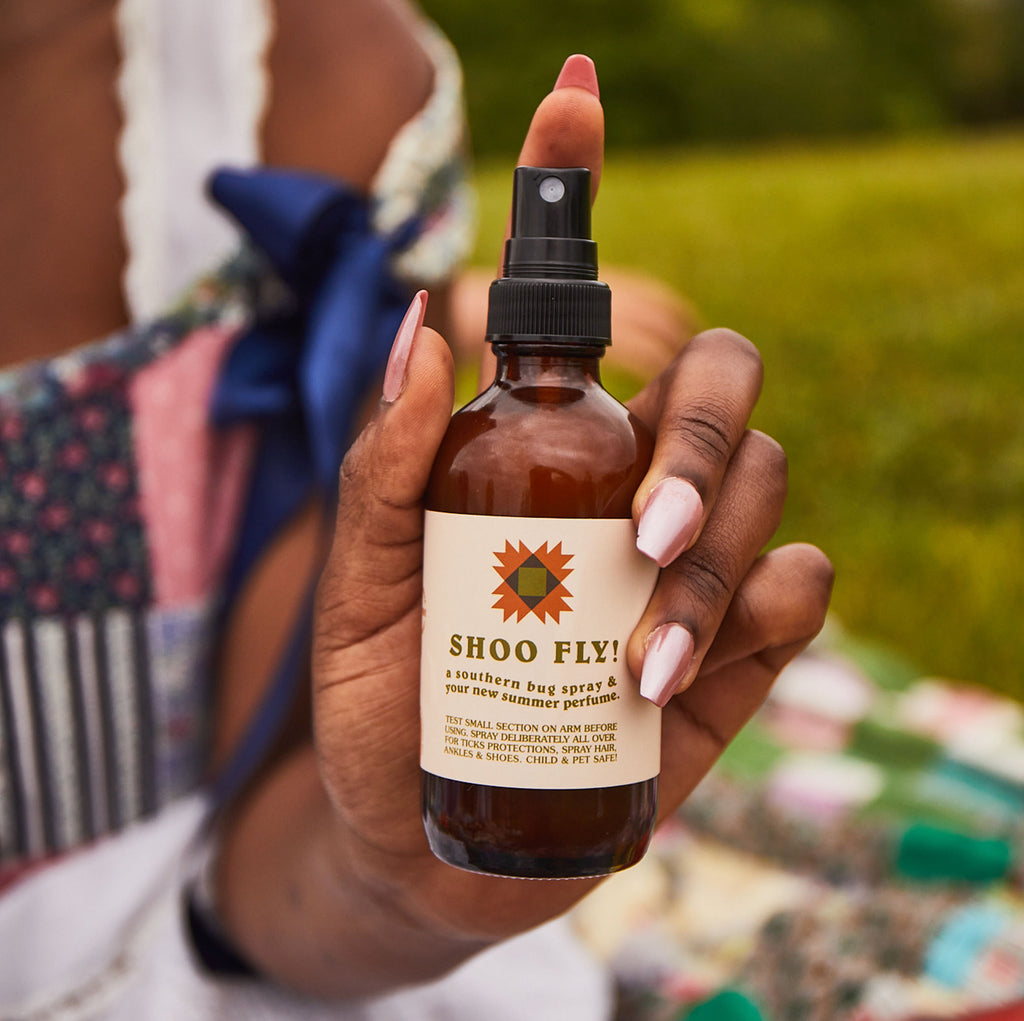 A woman holds a bottle of Shoo Fly! Southern bug spray and summer perfume while picnicking outdoors.
