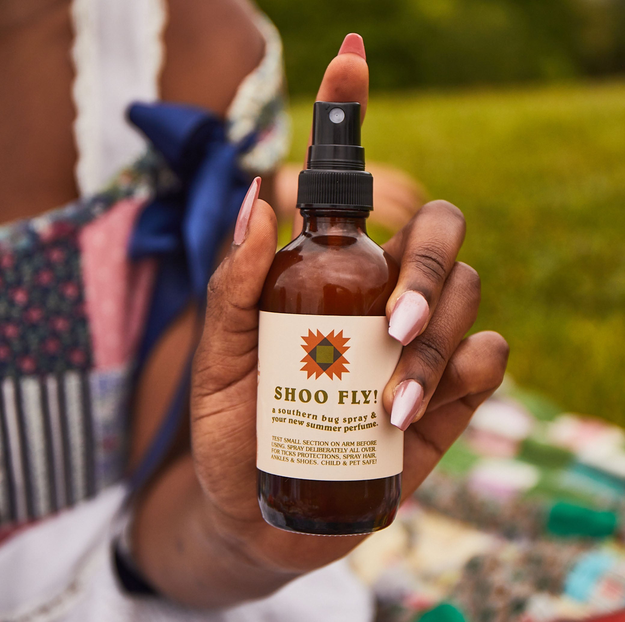 A woman holds a bottle of Shoo Fly! Southern bug spray and summer perfume while picnicking outdoors.
