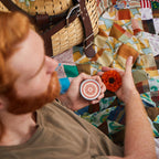 A red-haired person holds a "Redneck Redemption" flower compact and a marigold on a colorful patchwork quilt beside a woven picnic basket.
