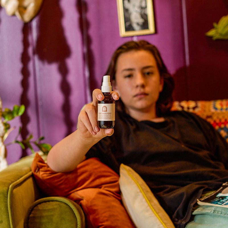 Person showcases sore throat spray while relaxing on a colorful couch at home.
