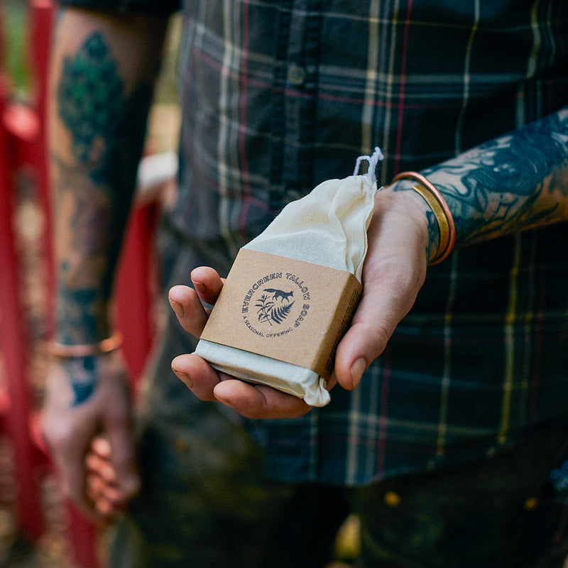 A tattooed person holds a wrapped bar of Evergreen Tallow Soap, showcasing its natural ingredients and seasonal offering.
