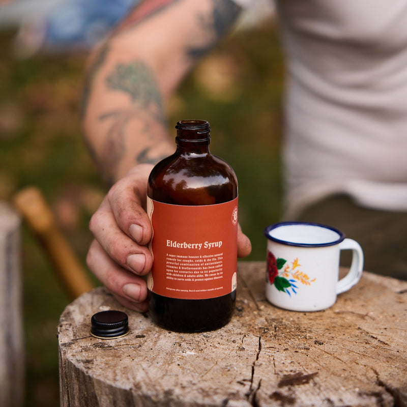 A tattooed arm holds a bottle of elderberry syrup next to a floral mug on a wooden stump.
