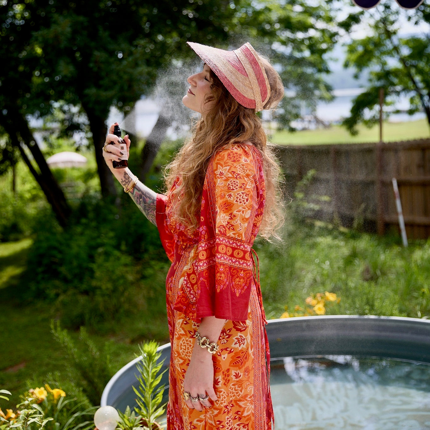 A woman in a bohemian dress and sun visor sprays a refreshing facial mist on a sunny summer day outdoors.
