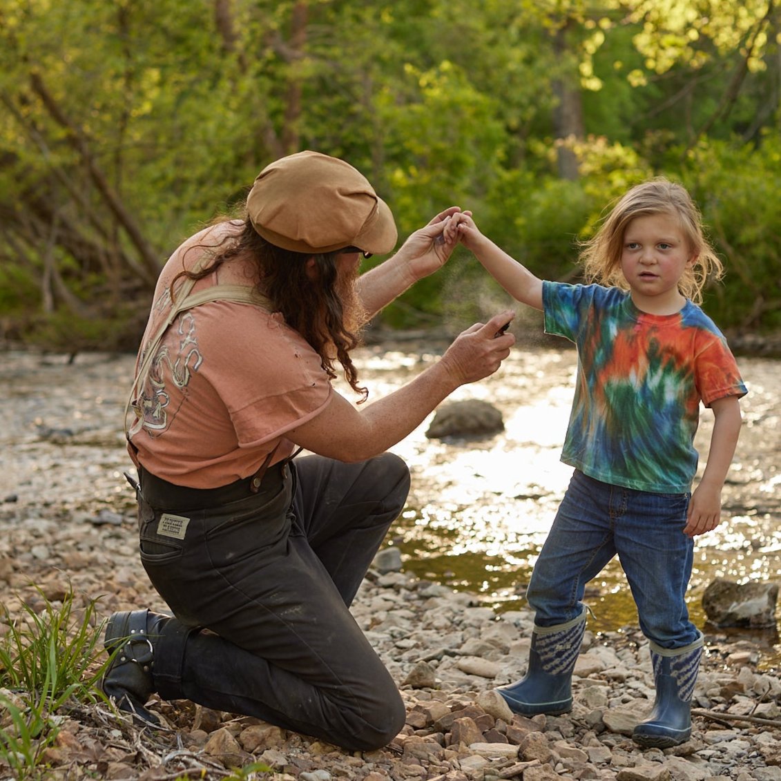 A parent sprays bug repellent on their child near a rocky creek bed in the woods.
