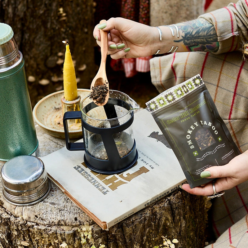Person prepares Cedar Hill Homestead Smokey Taboo loose leaf tea outdoors, using a wooden spoon to fill a glass press on a vintage Tennessee Hill Folk book.
