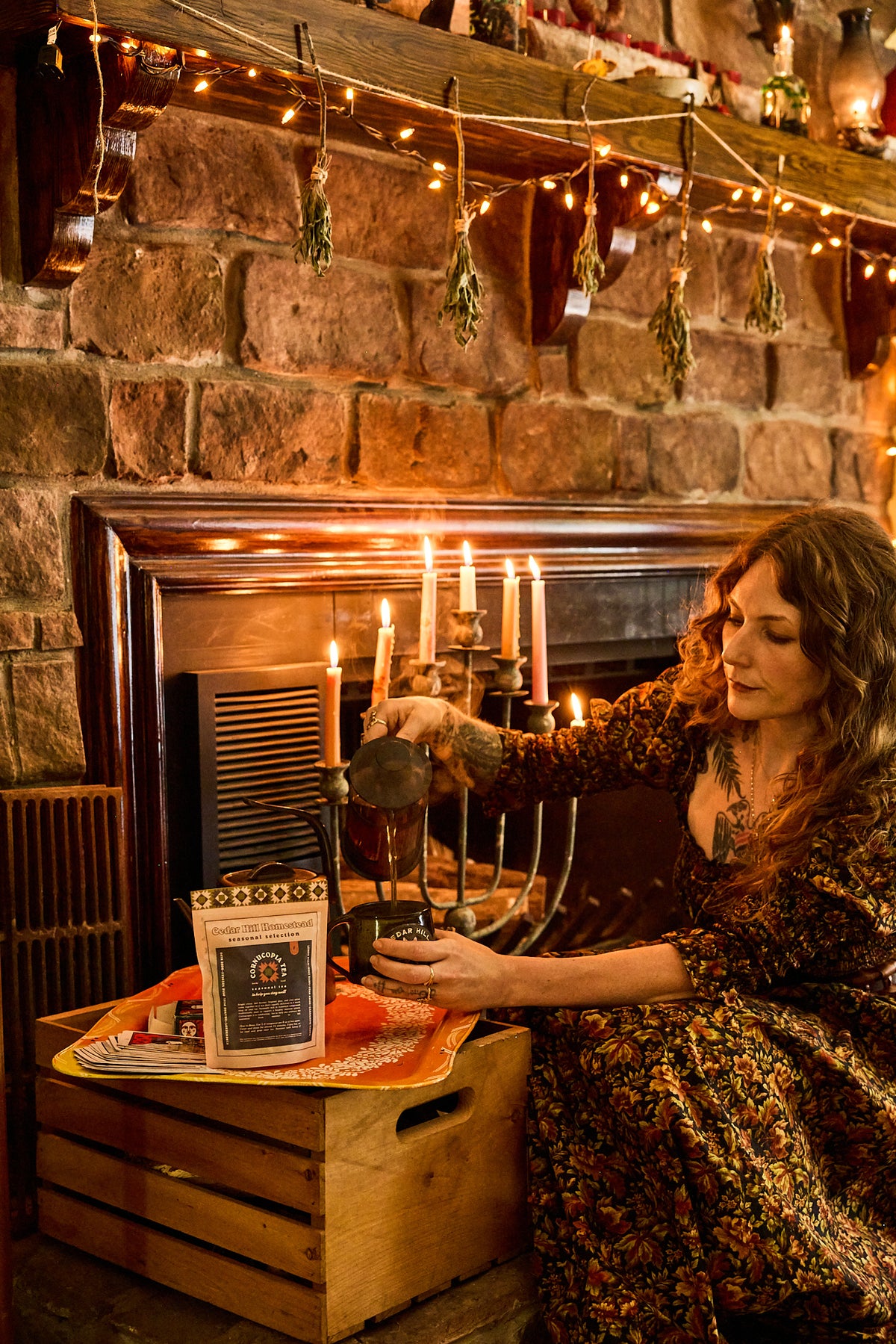 A woman in a floral dress stands by a rustic stone fireplace, pouring hot water from a metal kettle into a cup. A bag of coffee or tea and a small jar sit on a wooden crate in front of the fireplace, with candles and warm string lights visible in the background.
