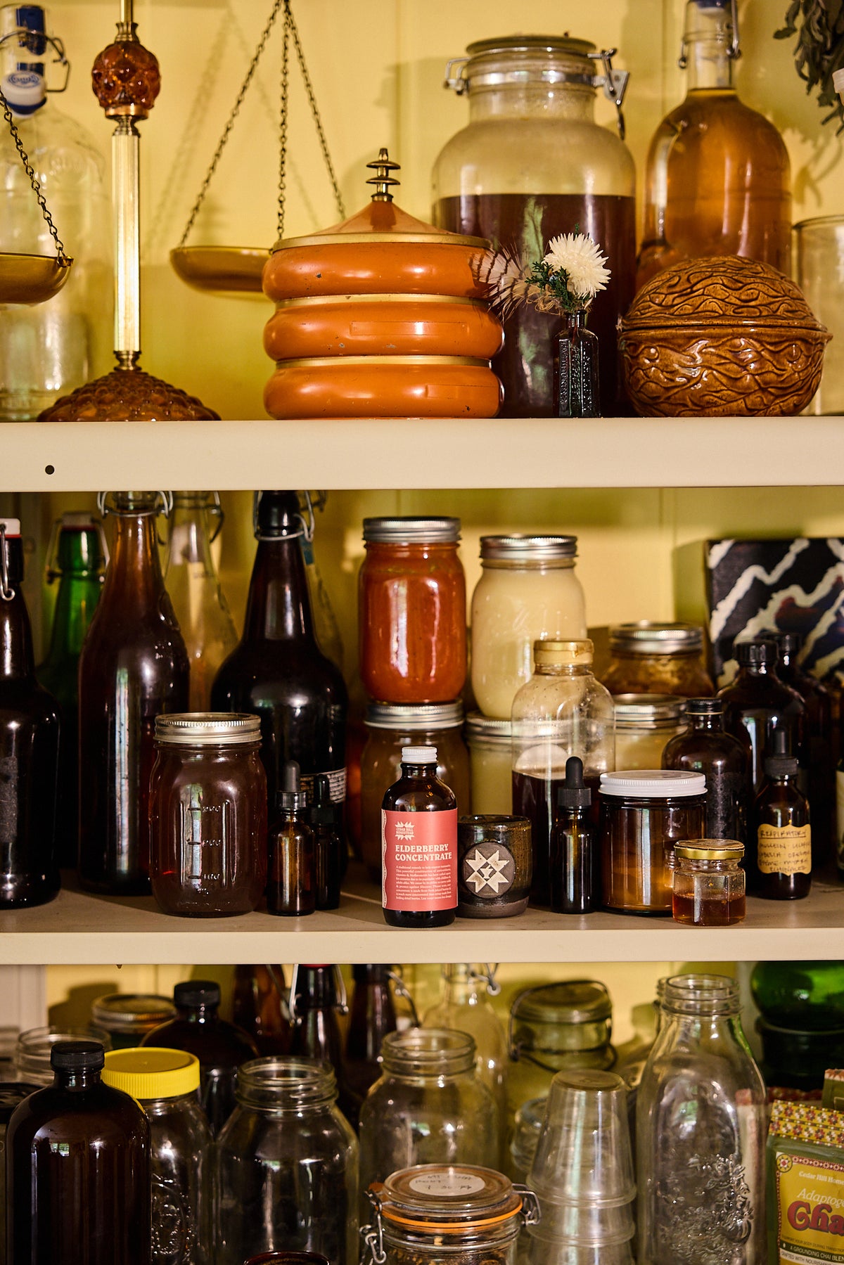 A collection of vintage and antique glass bottles, jars, and apothecary containers arranged on wooden shelves against a warm yellow background.