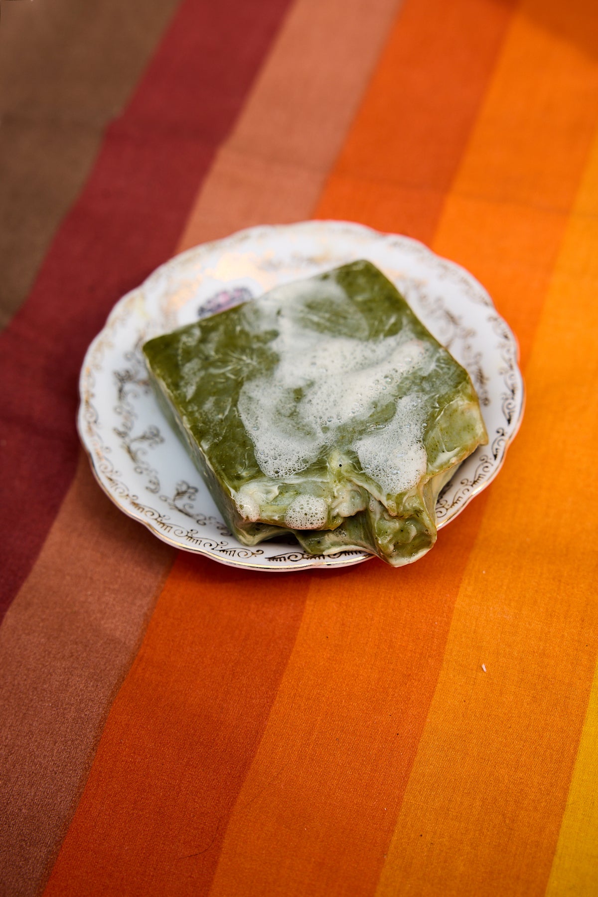 A square green food item, possibly a piece of layered cake or pastry with white frosting on top, served on a small white plate with a gold pattern, set against a striped orange, brown, and red tablecloth.