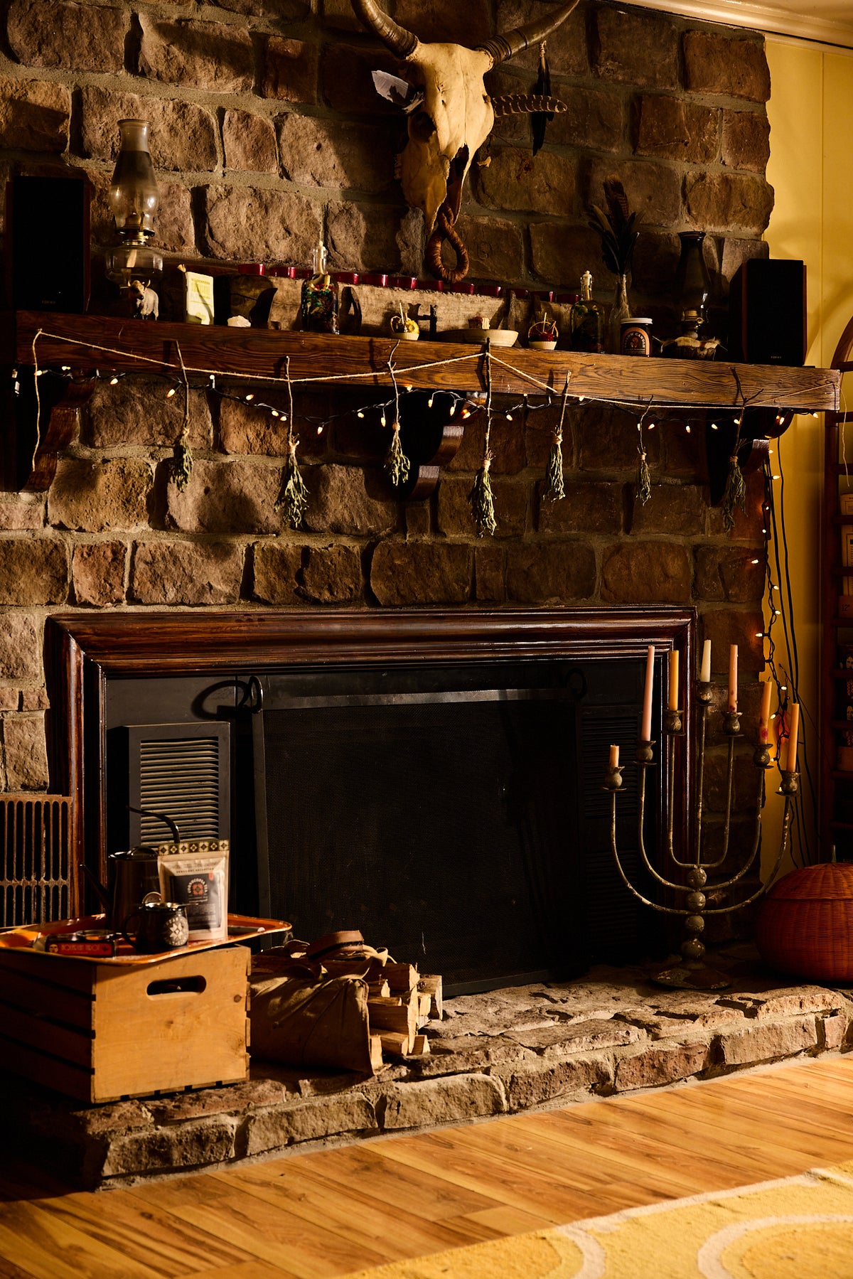 A rustic stone fireplace with a dark wood mantel decorated with candles, glassware, and hanging ornaments, set in a cozy room with a wooden floor and a moccasin on the hearth.