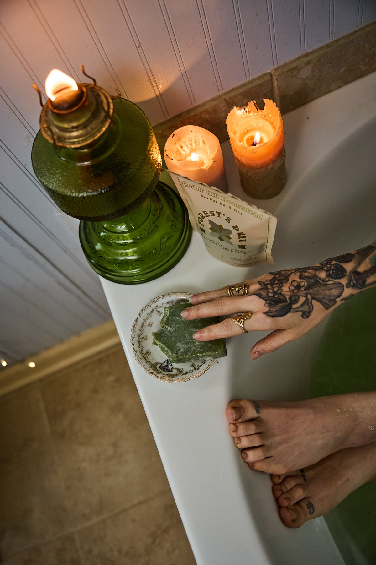A hand with a floral tattoo rests on a round glass dish containing green leaves, set on a white bathtub ledge alongside a green oil lamp, two lit candles, and a book. A person's bare feet are visible at the bottom right.