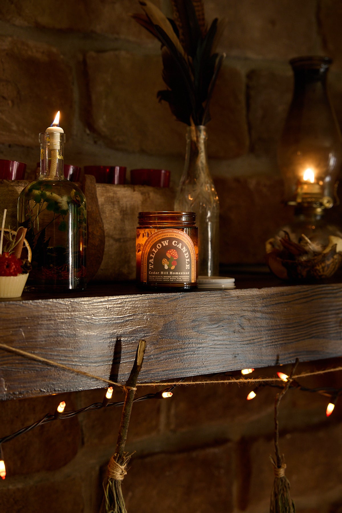 A rustic amber glass jar candle labeled 'Tallow Candle' sits on a dark wooden mantel in a cozy room setting with a stone fireplace, lit candles, and warm string lights.