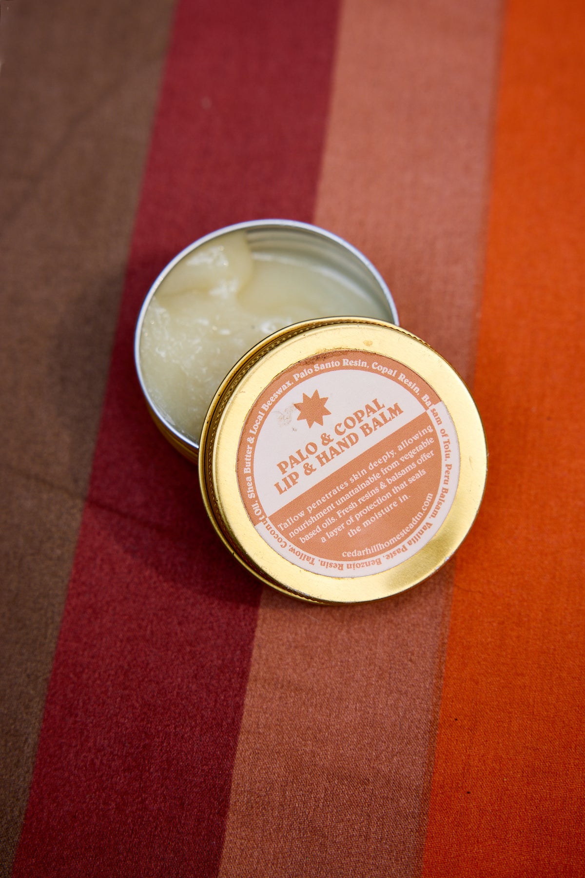 An open round metal tin of lip and hand balm with its lid placed beside it, showing the pale balm inside. The lid label reads 'Palo & Coalm Lip & Hand Balm.