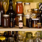 A small amber spray bottle labeled 'Cure Therapeutics' stands on a wooden shelf among larger glass jars and bottles containing various preserved foods and liquids.