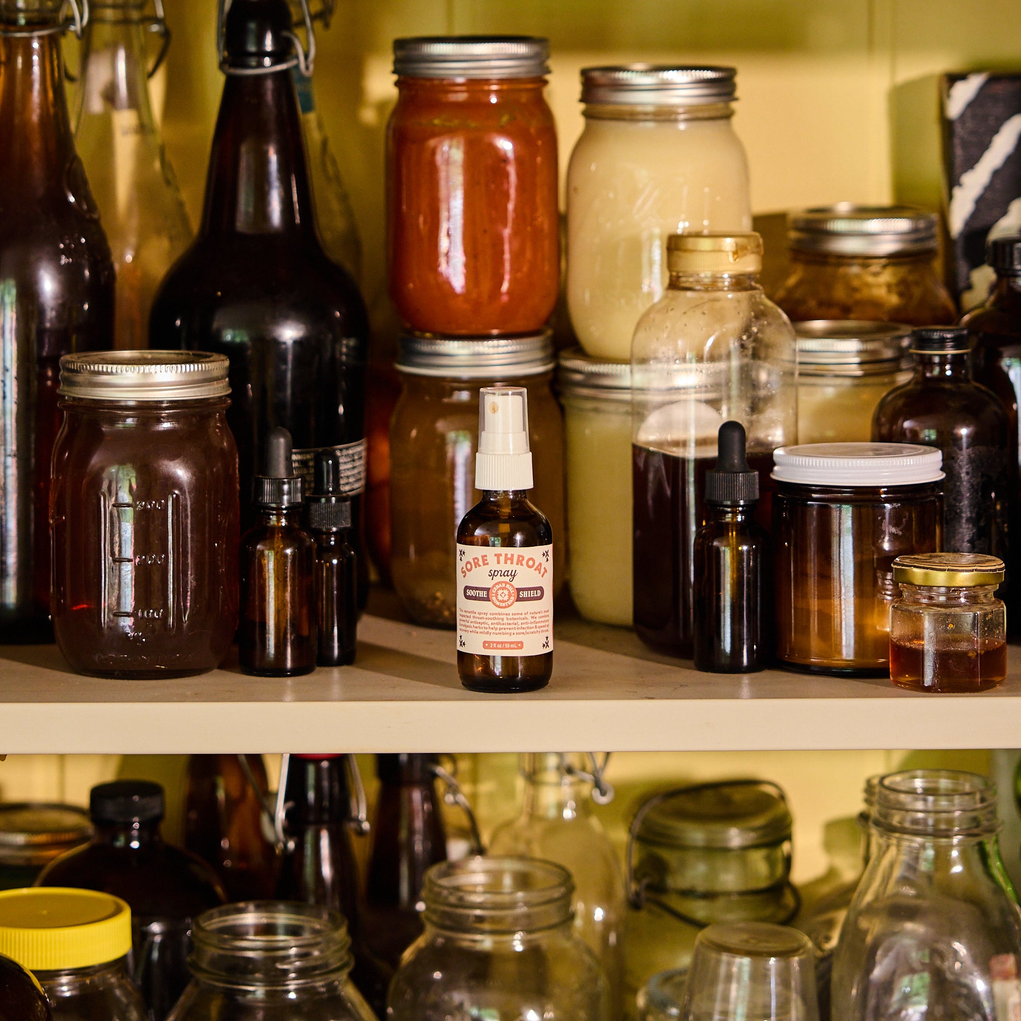 A small amber spray bottle labeled 'Cure Therapeutics' stands on a wooden shelf among larger glass jars and bottles containing various preserved foods and liquids.