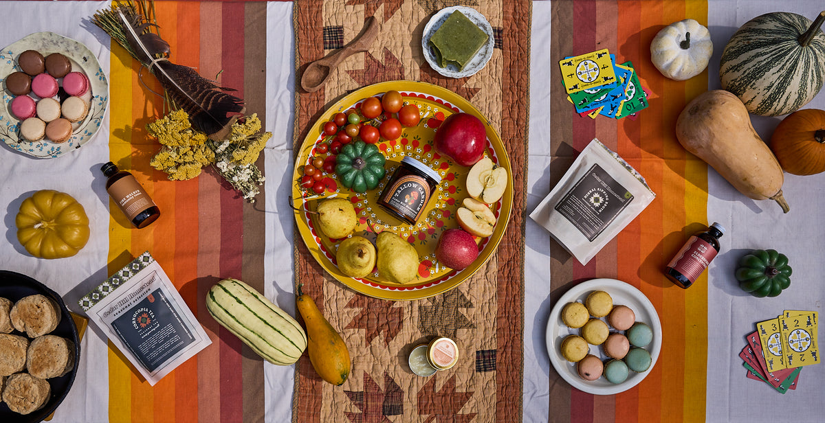 A styled flat-lay scene with a central round tray holding a jar and assorted fruits and vegetables, surrounded by autumn props including pumpkins, a striped cloth, books, cards, and small bottles on a wooden surface.