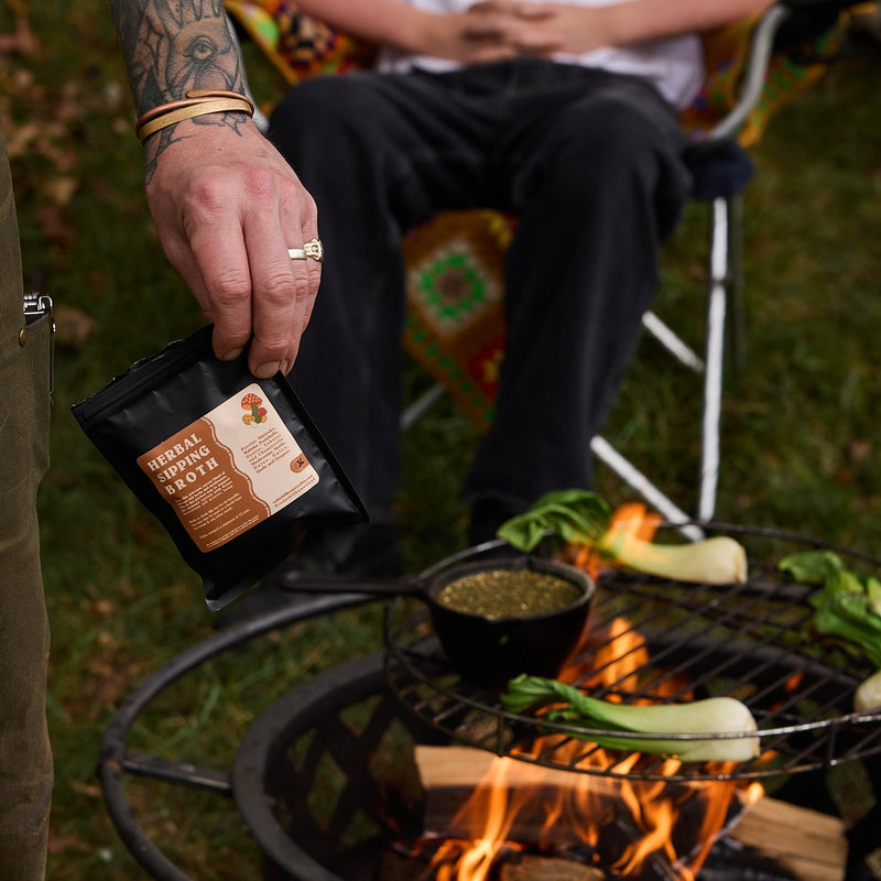 A tattooed man holds herbal sipping broth while preparing vegetables over an open fire outdoors.
