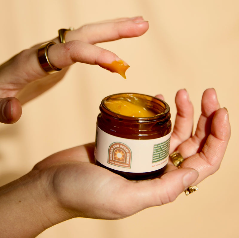 Hands apply Golden Sun Solid Serum from a brown glass jar.
