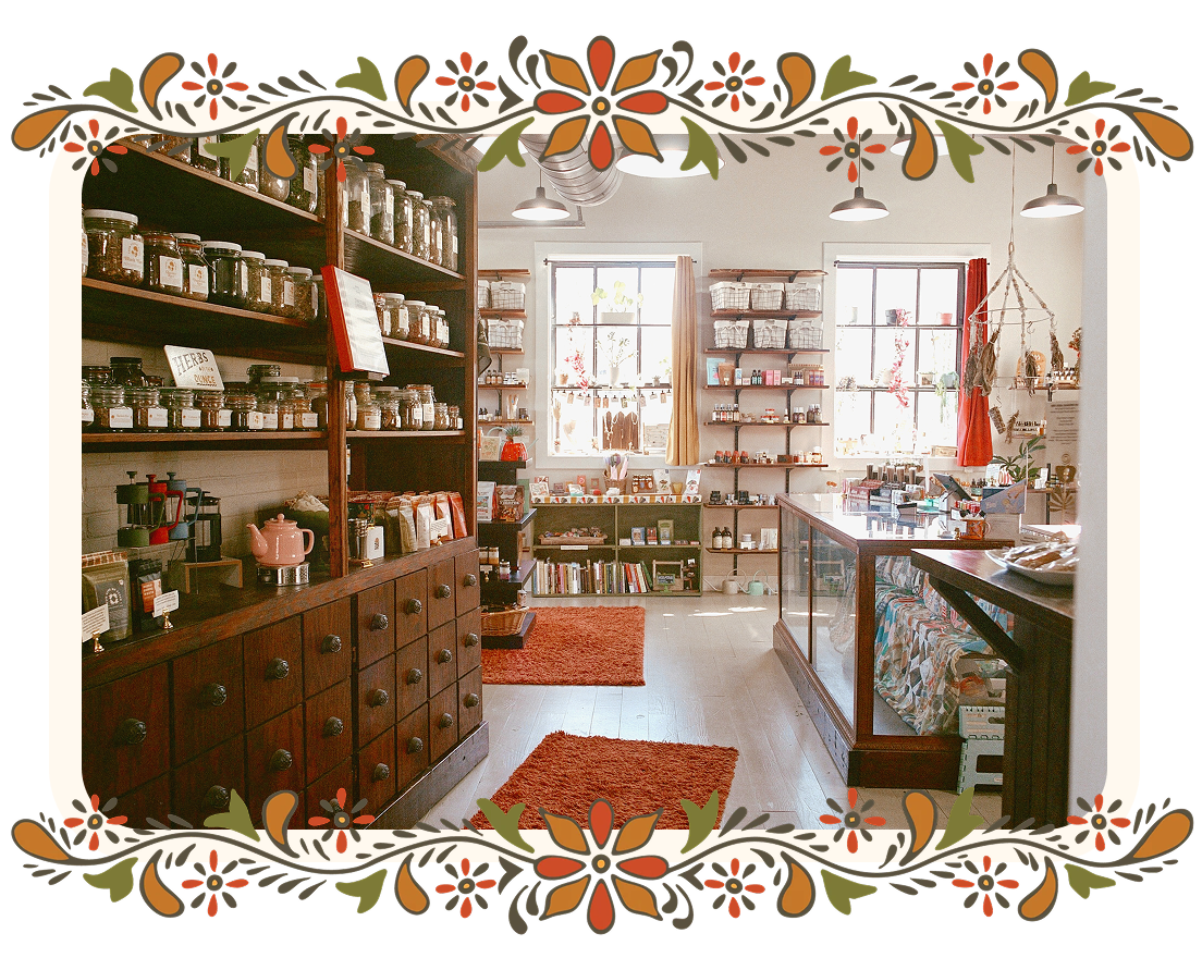 A cozy, rustic shop interior with wooden shelves filled with jars and bottles, a glass display counter, and warm orange-red rugs on a light floor. The photo is framed by a decorative floral border.