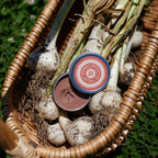 A woven basket holds freshly harvested garlic bulbs and open tin of Redneck Redemption bug bite relief balm, sitting outdoors in the grass.
