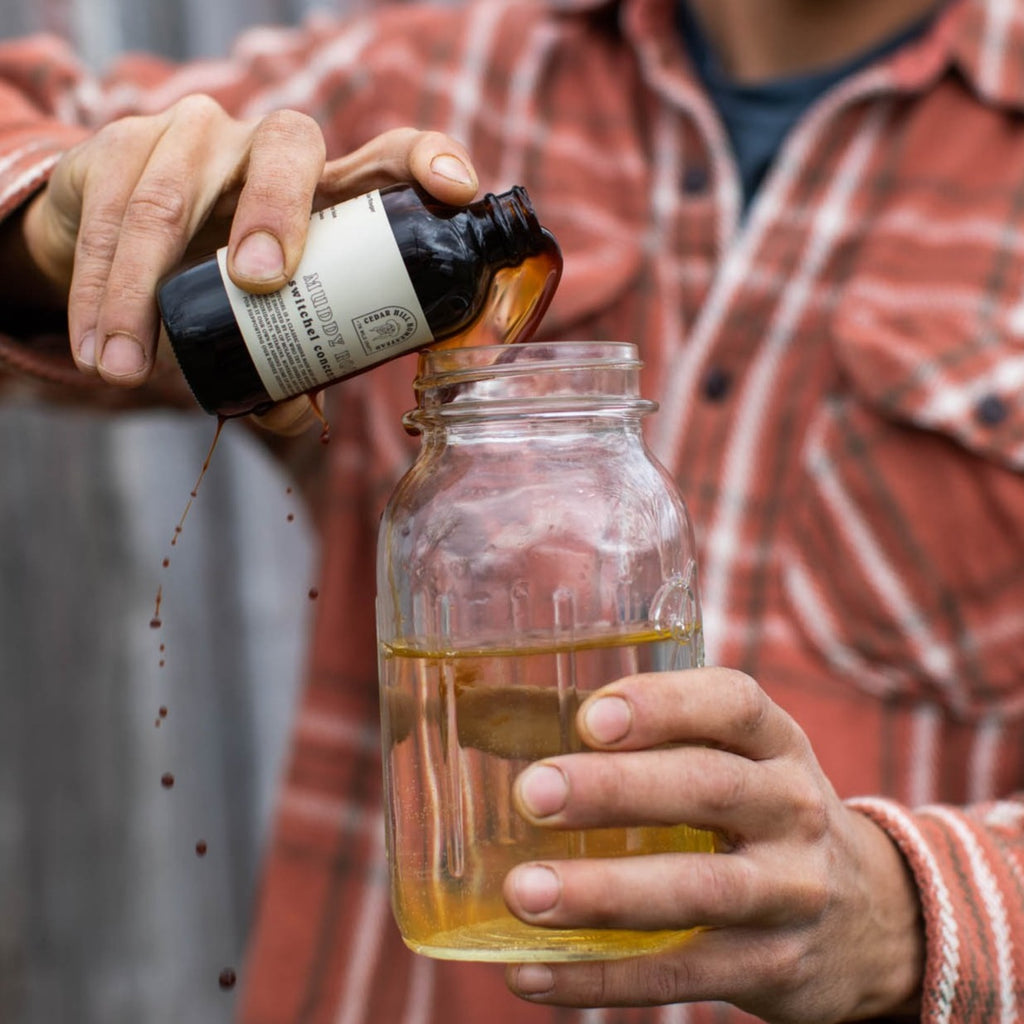 Someone wearing a plaid shirt pours Muddy Roar switchel concentrate from a dark bottle into a glass jar.
