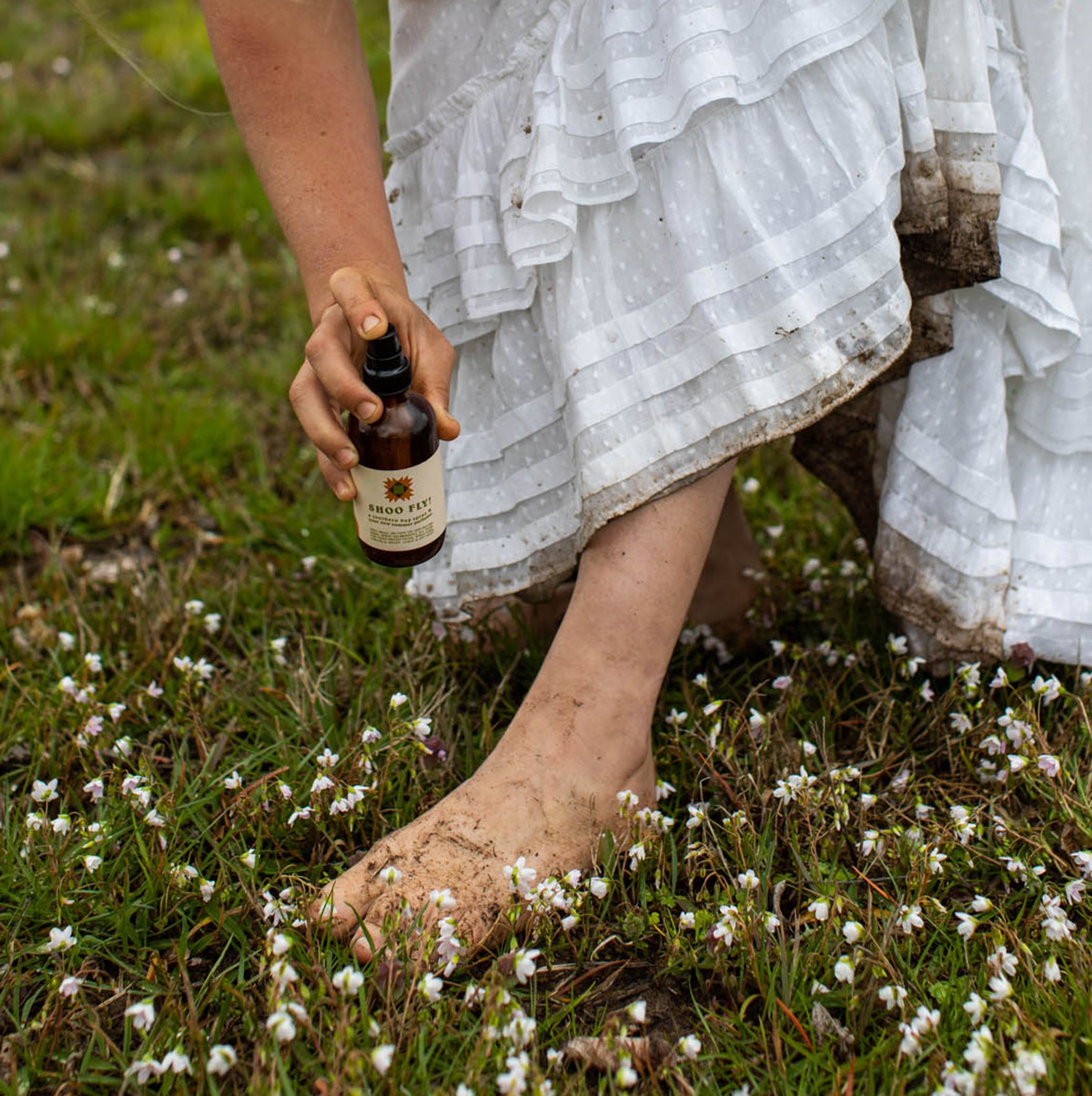 A barefoot woman in a mud-splattered white dress holds a bottle of Shoo Fly bug repellent outdoors amongst wildflowers.
