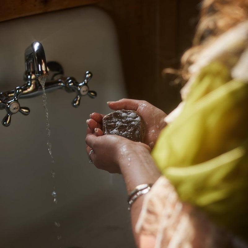A person washes their hands with a natural, dark-colored soap under a running chrome faucet in a bright sink.
