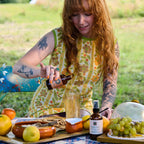 Woman at a picnic setting with food and drinks on a blanket in a park.
