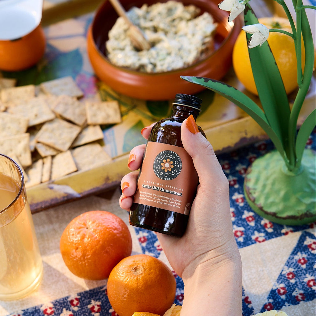 Hand holding a bottle of olive oil over a table with oranges, crackers, and a glass of juice.