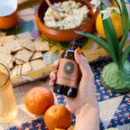 Hand holding a bottle of olive oil over a table with oranges, crackers, and a glass of juice.