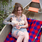 Woman with tattoos sitting on a boat covered with a red and blue checkered blanket, holding a bottle.
