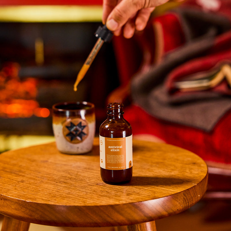A hand squeezes a dropper, adding cedarhillhomesteadtn.com antiviral elixir to a ceramic mug against a cozy fireside backdrop.
