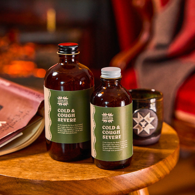 Two bottles of Cedar Hill Homestead Cold & Cough Severe sit on a wooden stool, offering natural cough relief.
