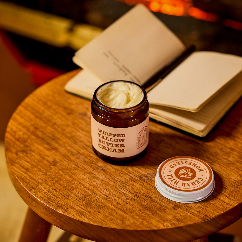 A jar of Cedar Hill Homestead whipped tallow butter cream sits open on a wooden stool beside a handwritten journal, creating a cozy, fireside skincare scene.
