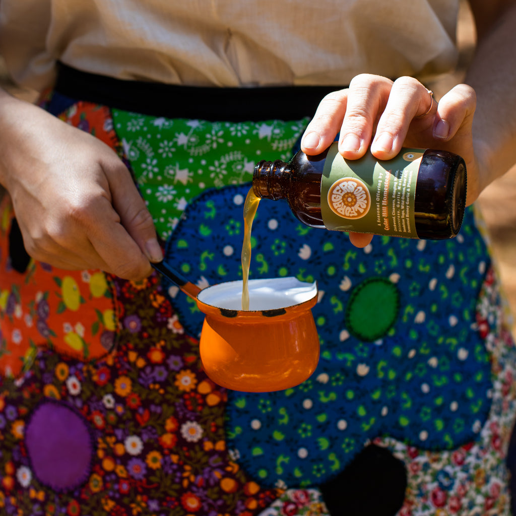 Woman wearing a floral apron pours Cedar Hill Homestead herbal infused oil into a small orange pot.
